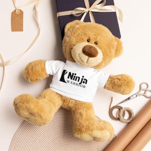 May include: A plush, tan teddy bear wearing a white t-shirt with the words "Ninja Warrior" in black. The bear is lying on a light surface, with a wrapped gift box, scissors, and craft supplies nearby. A gift tag hangs from a ribbon.