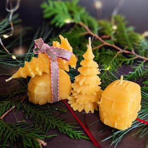 Op de afbeelding: Drie gele bijenwaskaarsen in de vorm van een camper, een kerstboom en een ingepakt cadeau. De kaarsen zijn versierd met een rood lint en een bruin en wit lint. Groene dennentakken omringen de kaarsen.