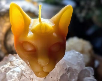 Puede incluir: Una vela amarilla con forma de gato con una mecha, colocada sobre un grupo de cristales de cuarzo transparente. La vela tiene una cara detallada con orejas grandes y un símbolo de ojo en la frente. El color de la vela pasa de amarillo a naranja.