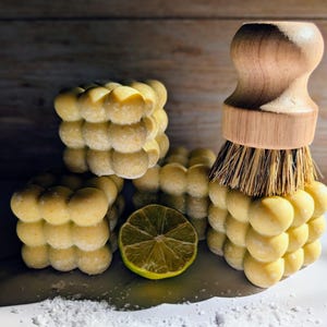 May include: A display of yellow, bubble-shaped soaps and a wooden-handled brush with bristles. A halved lime and scattered white salt are also present. The soaps are stacked, creating a textured, spa-like aesthetic.