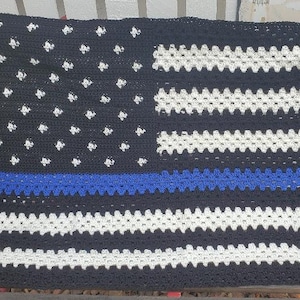 May include: A handmade crochet blanket featuring a design reminiscent of the American flag. The blanket has a black field with white stars, white and black stripes, and a blue stripe. The blanket is draped over a red chair.