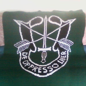 Op de afbeelding: Een groene gehaakte deken met een wit geborduurd embleem met een zwaard en pijlen. Het embleem is omgeven door een zwarte cirkel met de tekst "De Oppresso Liber" in wit.
