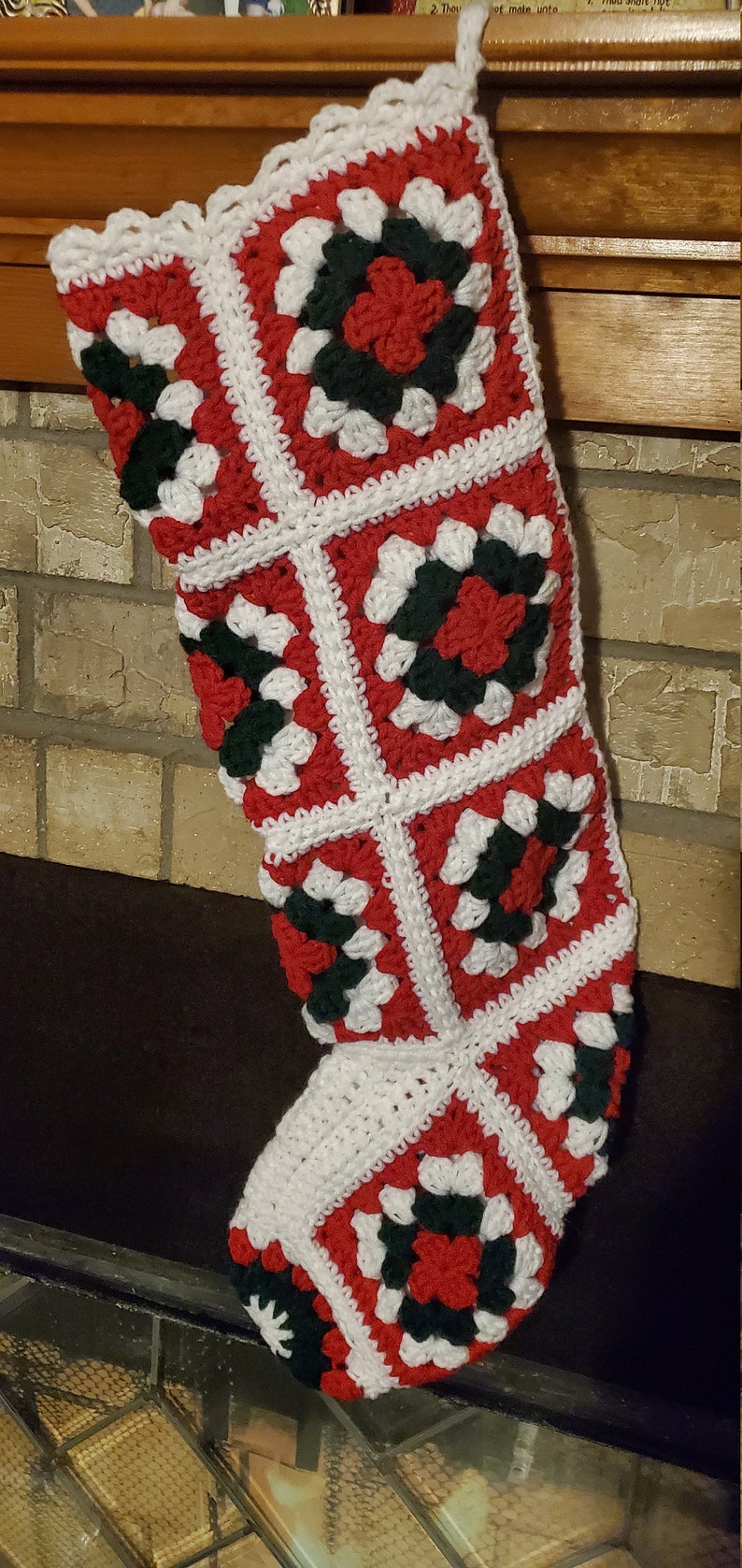 Pode incluir: Uma meia de Natal de croch&ecirc; com um esquema de cores vermelho, branco e verde. A meia &eacute; feita de quadrados com um desenho de flor no centro de cada quadrado.