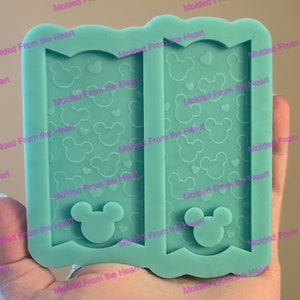 May include: A two-part silicone mold for making resin crafts. The mold features a repeating pattern of Mickey Mouse heads and hearts. The mold is light blue and has the text "Molded From the Heart" repeated around the edges.