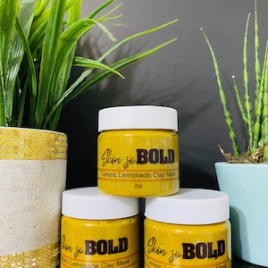 Peut inclure: Trois pots de 2 onces de masque d'argile à la citronnade au curcuma. Les pots sont étiquetés "Skin so BOLD" et sont empilés les uns sur les autres. Les pots sont sur une surface sombre avec des plantes vertes en arrière-plan.