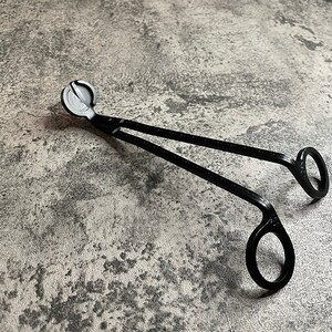 May include: Black metal candle wick trimmer with a round handle and two loops. The tool is designed to trim wicks on candles.