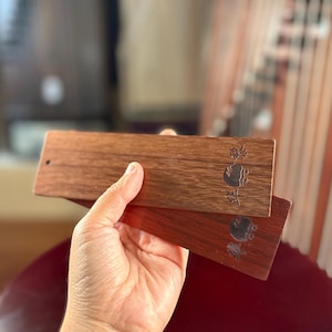 Può includere: Due ponti rettangolari in legno di cetra in varie tonalità di marrone sono tenuti in mano. Ogni ponte ha un piccolo foro e un disegno inciso con caratteri cinesi. La venatura del legno è visibile.