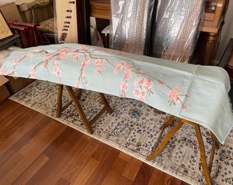 Op de afbeelding: Een lichtblauwe stoffen tafelrunner met een roze bloemenpatroon. De runner ligt over twee houten bokjes.