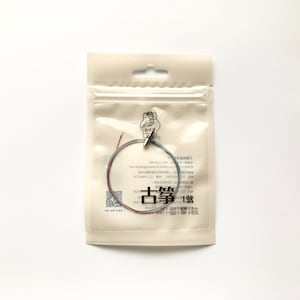 May include: A clear plastic bag containing a silver string with red and blue accents. The bag has a white label with Chinese text and a logo that says "古筝".