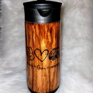 May include: A brown and black stainless steel travel mug with a wood grain pattern. It has a black lid and a white design that says "Peace Love Camping" with a peace sign, heart, and a camper van.