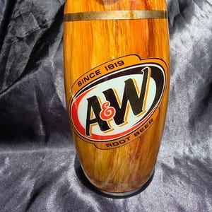 May include: A cylindrical tumbler with a wood grain design and a clear lid. The A&W Root Beer logo is prominently displayed on the front, with the words "Since 1919" and "Root Beer" included in the design. The tumbler has a gold-colored band around the middle.