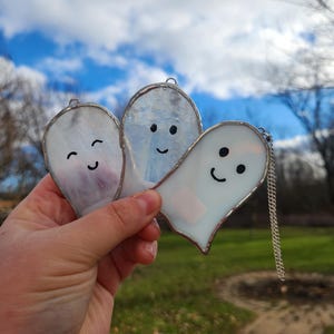 以下が含まれることがあります： 笑顔の顔をした、手作りのステンドグラスのゴーストオーナメント3つ。各ゴーストは銀色で縁取られ、吊り下げ用の小さなループが付いています。1つのゴーストにはチェーンが付いています。ゴーストは白、紫、青の色合いです。