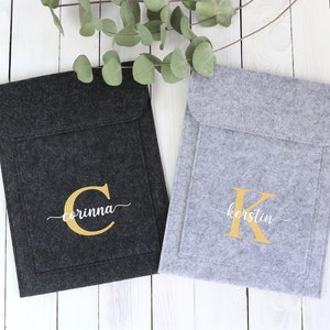 May include: Two felt laptop sleeves with personalized monograms. The sleeve on the left is black with a gold "C" and the name "corinna" in white script. The sleeve on the right is gray with a gold "K" and the name "kerstin" in white script.