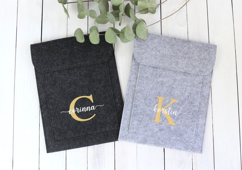 May include: Two felt laptop sleeves with personalized monograms. The sleeve on the left is black with a gold "C" and the name "corinna" in white script. The sleeve on the right is gray with a gold "K" and the name "kerstin" in white script.