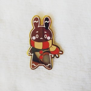 May include: Gold-coloured enamel pin featuring a gingerbread bunny wearing a red and yellow scarf. The bunny has brown eyes and a brown nose.
