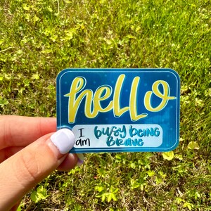 May include: A rectangular sticker with a teal background and rounded corners. The sticker features the word "hello" in yellow script, with the phrase "I am busy being brave" in a smaller font.