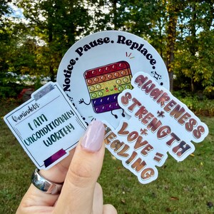 May include: A collection of stickers with positive affirmations and designs. One sticker features a colorful pop-it toy with the words "Notice. Pause. Replace." Another says "I AM UNCONDITIONALLY WORTHY." A third sticker reads "AWARENESS + STRENGTH = LOVE + HEALING."