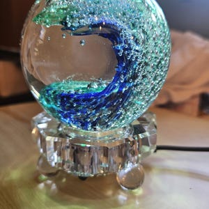 May include: A glass paperweight with a blue and green wave design inside. The paperweight is on a clear glass base with three legs.