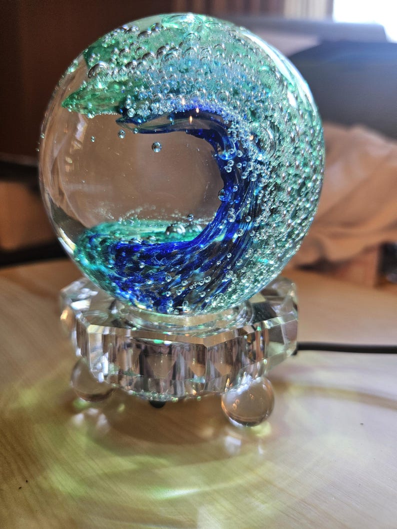 May include: A glass paperweight with a blue and green wave design inside. The paperweight is on a clear glass base with three legs.