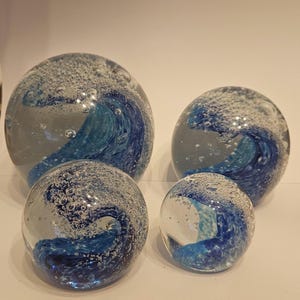 Könnte beinhalten: Vier klare Glaskugeln mit blauen Wellenmustern. Die Kugeln variieren in der Größe, wobei die größte einen Durchmesser von etwa 13 cm hat. Die Wellenmuster sind im Glas verwirbelt und erzeugen einen dynamischen, ozeanartigen Effekt. Blasen sind im Glas eingeschlossen.
