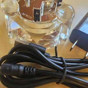 May include: A silver and clear glass electric lighter with a black cord and plug. The lighter has a clear glass base with four clear glass spheres on the top. The lighter has a silver metal top with a small silver tube on the top. The lighter is plugged into a black cord with a black plug.