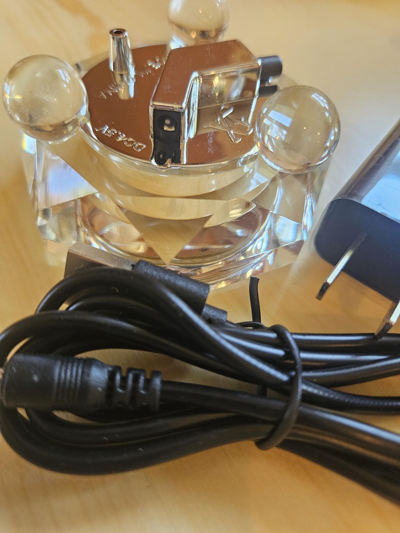 May include: A silver and clear glass electric lighter with a black cord and plug. The lighter has a clear glass base with four clear glass spheres on the top. The lighter has a silver metal top with a small silver tube on the top. The lighter is plugged into a black cord with a black plug.