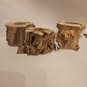 Puede incluir: Tres candelabros rústicos de madera. Cada soporte está hecho de una sección de tronco de árbol, mostrando la veta y textura natural de la madera. Los soportes tienen una hendidura circular en la parte superior para velas.