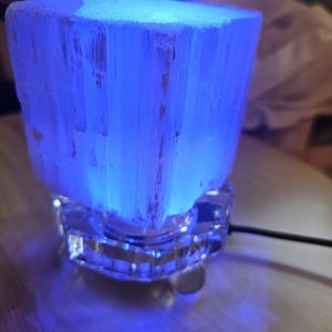 May include: A white selenite crystal lamp with a blue light, sitting on a clear glass base. The lamp is plugged in and the cord is visible.