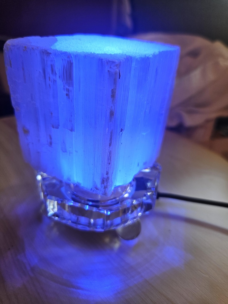 May include: A white selenite crystal lamp with a blue light, sitting on a clear glass base. The lamp is plugged in and the cord is visible.