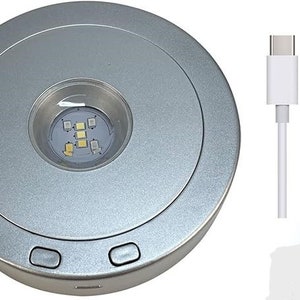 Puede incluir: Una plataforma de carga redonda plateada con un cable USB-C blanco y un cable USB-A blanco. La plataforma de carga tiene una pequeña luz LED circular en el centro.