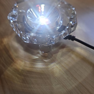 May include: A clear crystal base with a small, bright white LED light in the center. The base has a faceted design and a clear glass stem.