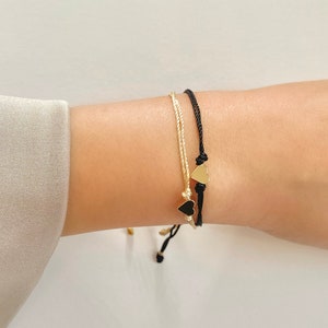 以下が含まれることがあります： ハート型のチャームが付いた2つのブレスレット。1つは黒いコードと金のハートチャーム、もう1つはクリーム色のコードと金のハートチャームが付いています。ブレスレットは手首に着用されています。