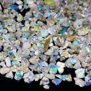 Könnte beinhalten: Eine Ansammlung roher, unpolierter Opalsteine mit unregelmäßigen Formen und rauen Texturen. Die Steine zeigen eine Vielzahl schillernder Blitze in Blau-, Grün-, Gelb- und Weißtönen vor einer dunklen, neutralen und felsigen Matrix.