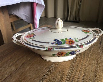 May include: A white ceramic serving dish with a floral design in shades of pink, blue, and yellow. The dish has a gold trim and a lid with a knob.
