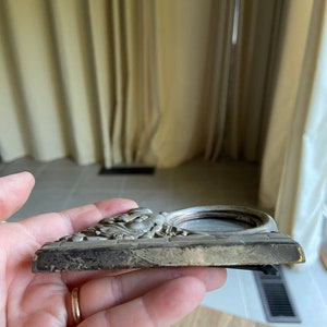 May include: A silver-toned picture frame with an ornate design featuring a raised, detailed image of a woman's face. The frame has a circular opening for a photo.