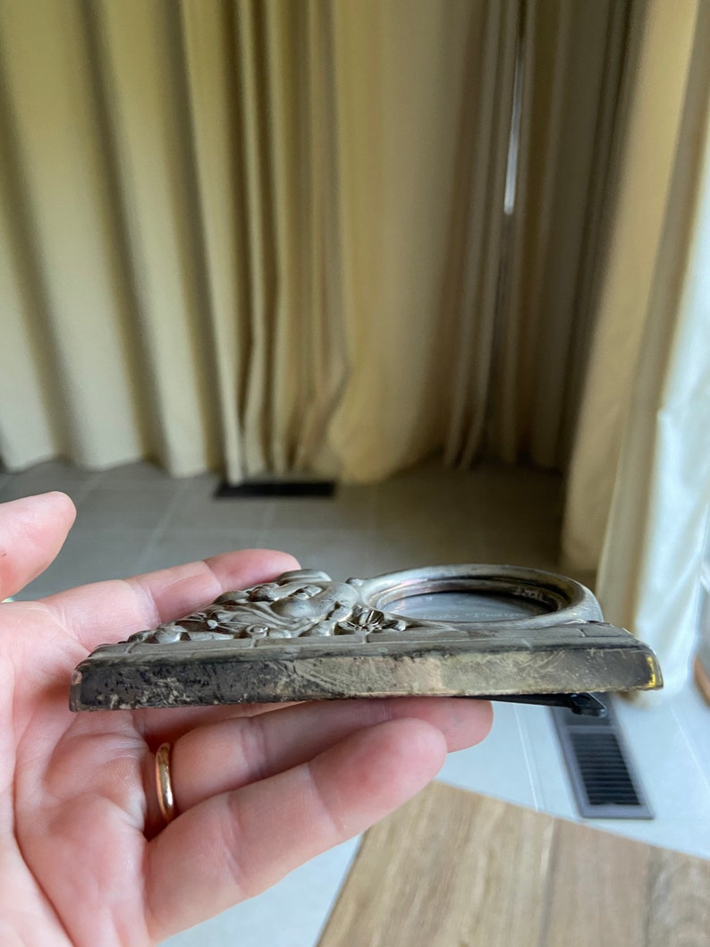 May include: A silver-toned picture frame with an ornate design featuring a raised, detailed image of a woman's face. The frame has a circular opening for a photo.