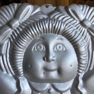 May include: A silver metal cake pan shaped like a doll with pigtails and a bow. The doll has a round face with large eyes, a small nose, and a wide smile.