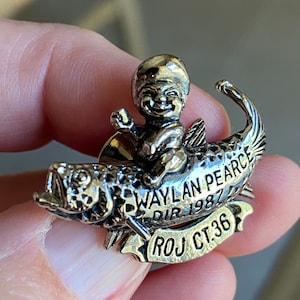 May include: Silver pin featuring a cartoon figure riding a fish. The fish has a banner that reads "Waylan Pearce Dir 1987 ROJ CT 36".