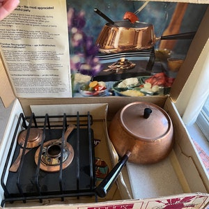May include: A vintage fondue set in its original box. The set includes a copper pot, a black metal burner, and a black handle. The box is brown with a picture of the fondue set on the front. The box also has the text "Fondue Bourguignonne - ein kulinarisches Gesellschaftsspiel" on the front.
