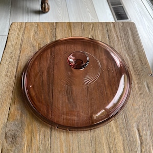 May include: A clear glass lid with a pink tint and a raised knob in the center. The lid is sitting on a wooden surface.