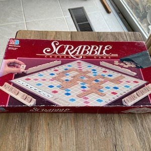 May include: A red and white box for the Scrabble board game. The box shows the game board with tiles placed on it, and the text "Scrabble Crossword Game" is printed on the box.