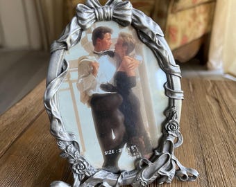 May include: Ornate silver-colored picture frame with a romantic image of a couple. The frame has a bow at the top and floral accents. The photo size is 3 x 5 inches.