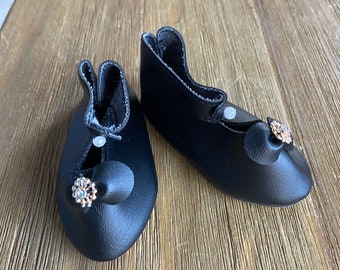 May include: A pair of black leather doll shoes with a small gold and white jeweled embellishment on each shoe.