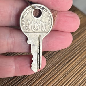 May include: A vintage silver-colored key with the word "Master" in a stylized font. The key has a circular head with the words "Milwaukee, Wis. U.S.A." around the edge. The key is held by a person.