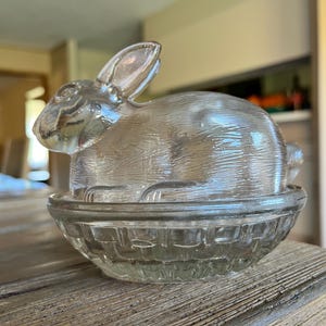 May include: A clear glass bunny-shaped dish with a basket weave base. The bunny sits atop the dish, with its ears perked up. The dish is made of clear glass and is likely used for serving or decoration.