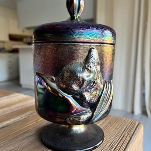 May include: Iridescent glass jar with a lid, featuring a raised relief of a sleeping fox. The jar has a textured surface and a pedestal base. The colors shift from purple to gold and green.