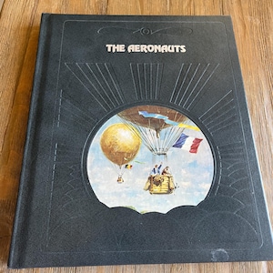 May include: A hardcover book titled "THE AERONAUTS" with a black cover. The cover features a circular illustration of hot air balloons and people in a basket. The illustration includes a French flag and a gold-colored balloon.