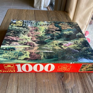 May include: A red and white box for a 1000 piece jigsaw puzzle featuring a landscape scene with a lake and trees. The box has the words "Golden Guild" and "Fully Interlocking" printed on it.