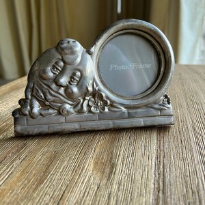 May include: Silver photo frame with a whimsical design featuring a cartoon face and flowers. The frame has a round opening for a photo and the words "Photo Frame" are printed on the glass.