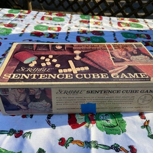 May include: A vintage Scrabble Sentence Cube Game box with a brown and red design. The box features a picture of people playing the game and the text "SCRABBLE SENTENCE CUBE GAME".